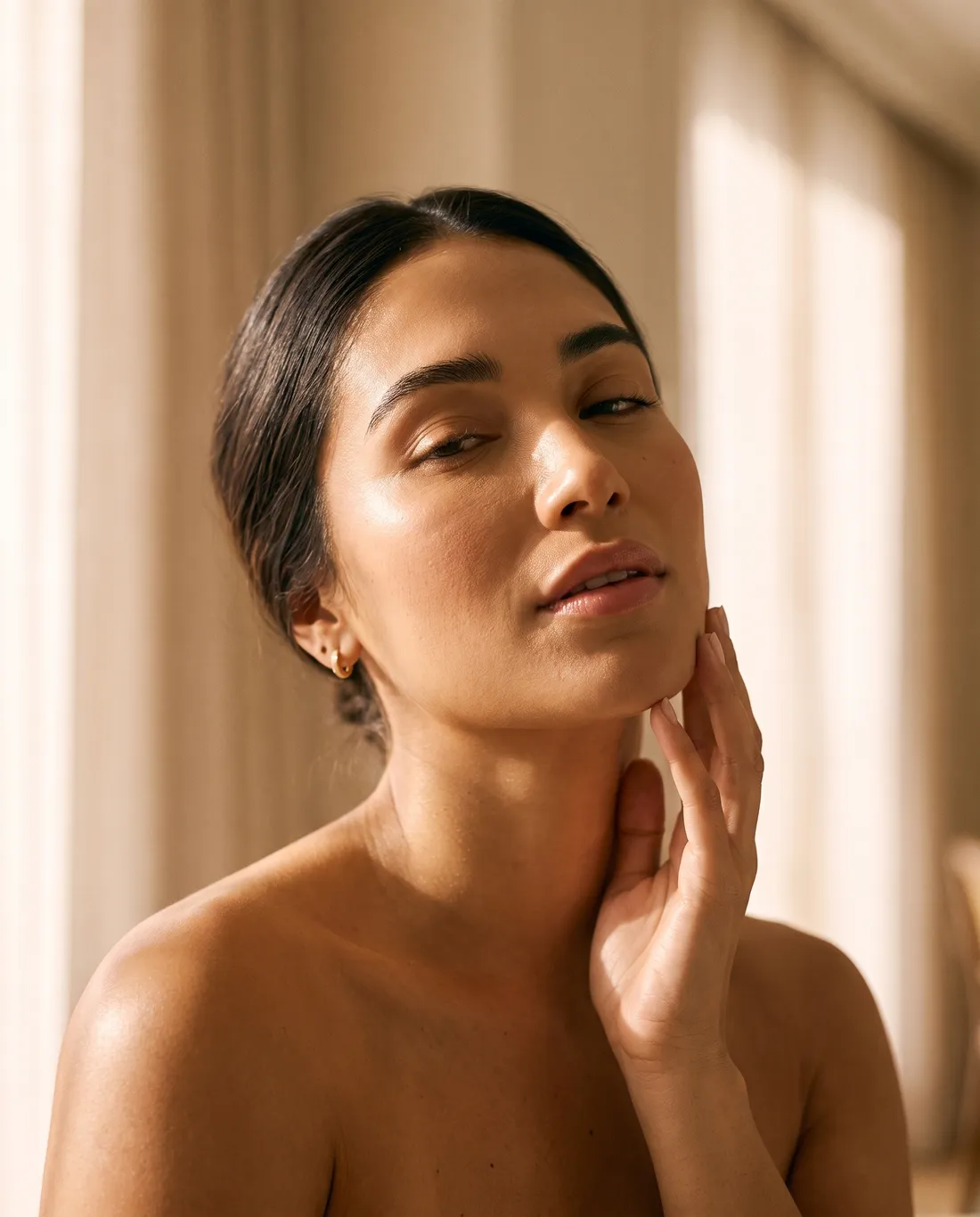 A woman with glowing skin resting her fingertips along her jawline in golden light
