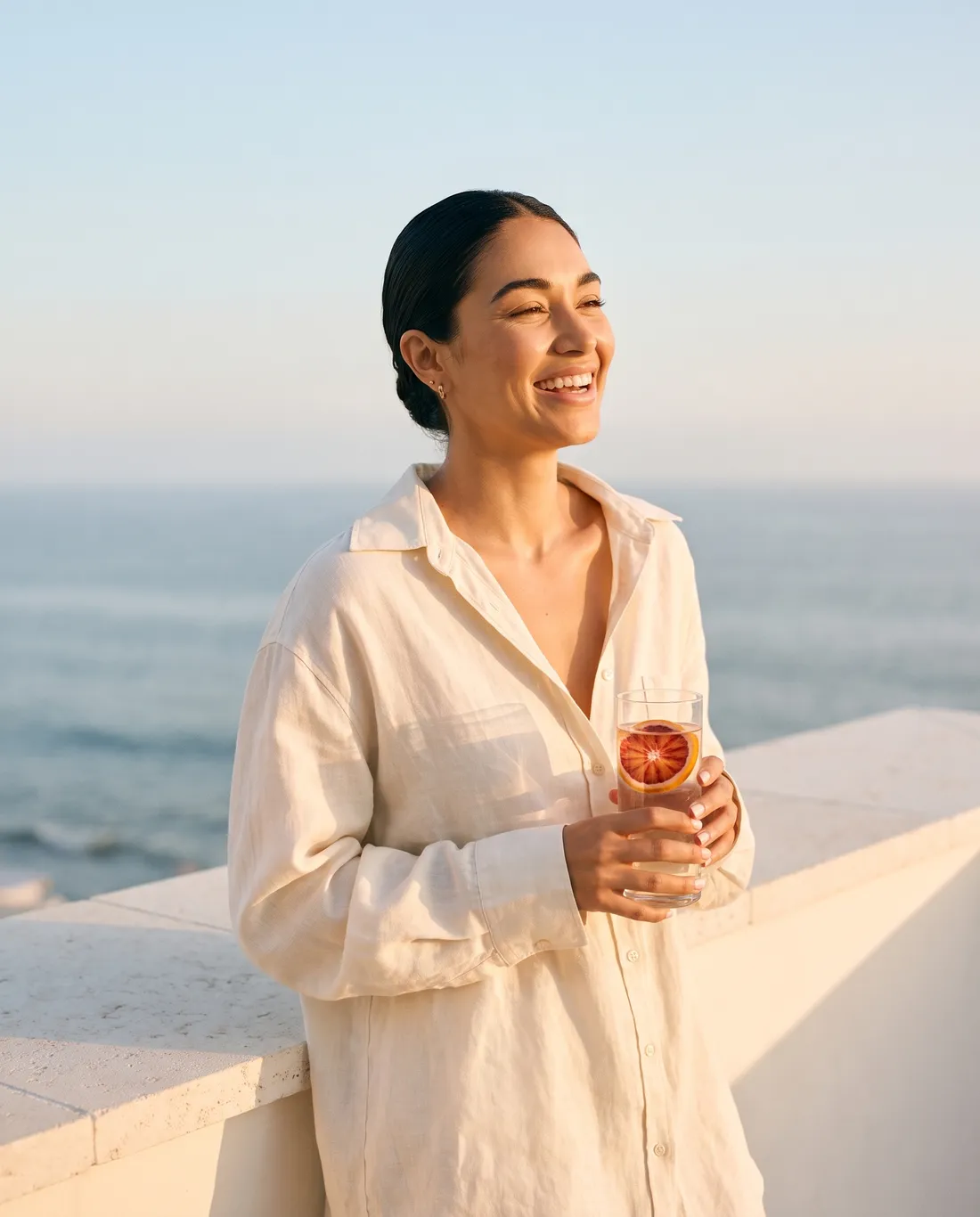 A laughing woman in a linen shirt on a sunlit terrace above the ocean, holding a glass of water